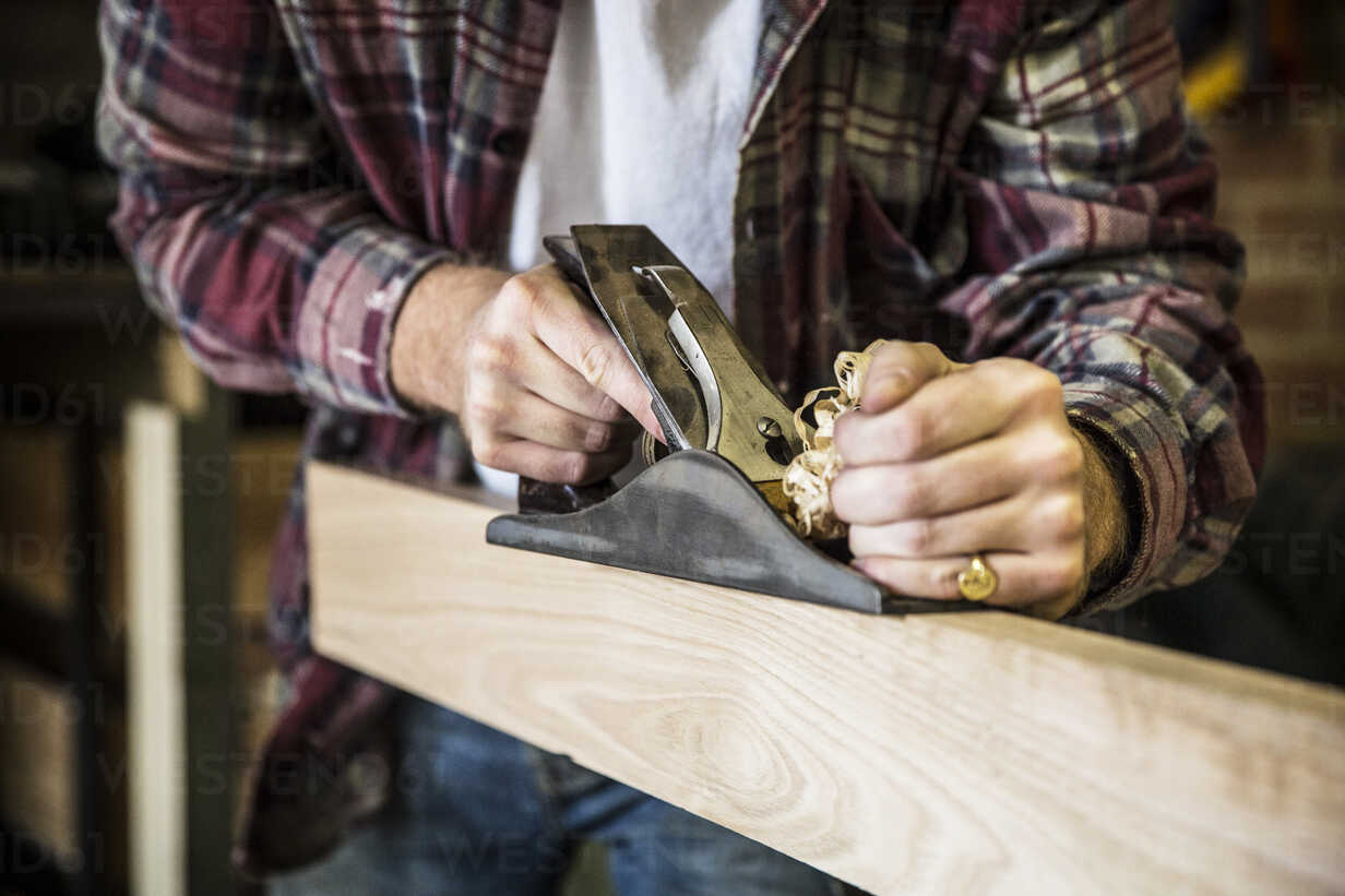 Close up of man working standing in a woodworking workshop, using plane on plank of wood.